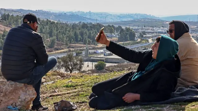 Three men sit on a grassy hill overlooking a prison. One man is lifting an arm with a phone in his hand, as if he's taking a selfie