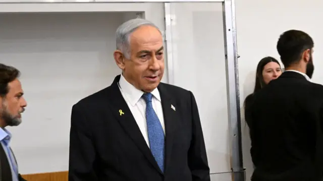 Netanyahu standing inside a room in a black suit, white shirt and blue tie. A yellow ribbon is pinned to his right lapel, a Israeli flag on his right. Two men and a woman also in the picture walking around