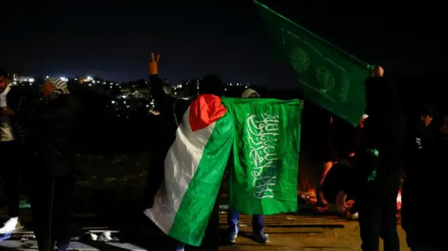 Two people at the centre of the image draped in a Palestinian flag (L) and a Hamas flag (R - green with white Shahada writing in white)