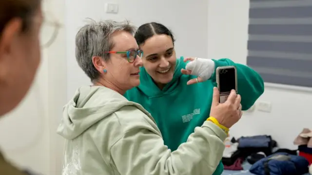 Emily Damari with her mother, Mandy. Emily is wearing a green jumper as she smiles at a phone during a phone call. Her mother is in a pastel green jumper holding the phone, they're in a white room with several objects on the ground