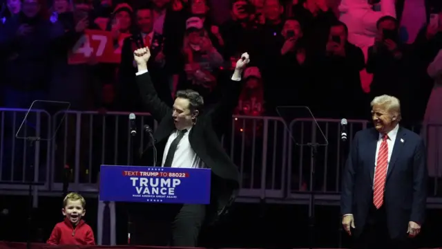 Musk holds his hands high alongside his son and Donald Trump on stage