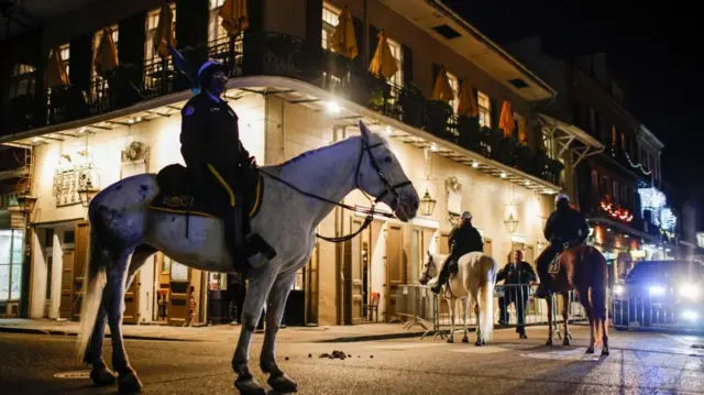 Three officers sit on horses at the corner of a street. Temporary metal gates have been set up across the road. It's night time and lights on a building and headlights can be seen