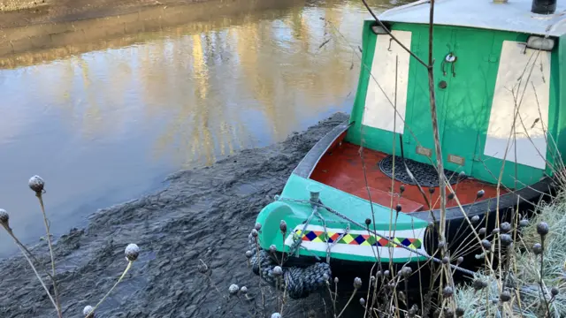 A green narrowboat stands on mud alongside the vastly-reduced water in the Bridgewater Canal