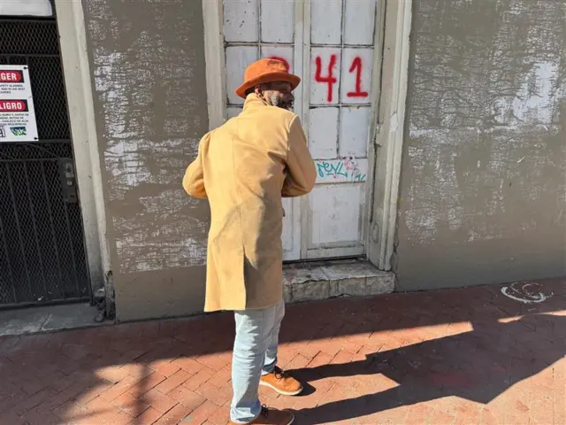 A man wearing a yellow jacket, turns to show the back of his jacket to the camera, with a building an door in the background and graffiti of the numerals 41 in red