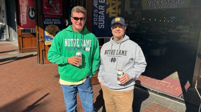 Two men smile at the camera. One is wearing a green jumper, holding a canned drink, with glasses and a chain. The other is wearing a grey cap, and grey jumper, brown trousers holding a canned drink.