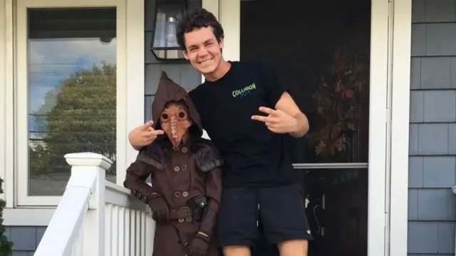 Matthew Tenedorio posing with a child in a what appears plague doctor mask and hood costume, Tenedorio is smiling and displaying two peace signs with his hands (extending his index ring fingers)