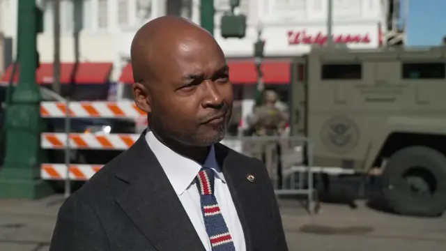 Jason Williams appears in black suit, white shirt and blue and red striped tie. He stands on the street outside in front of a law enforcement truck and building.