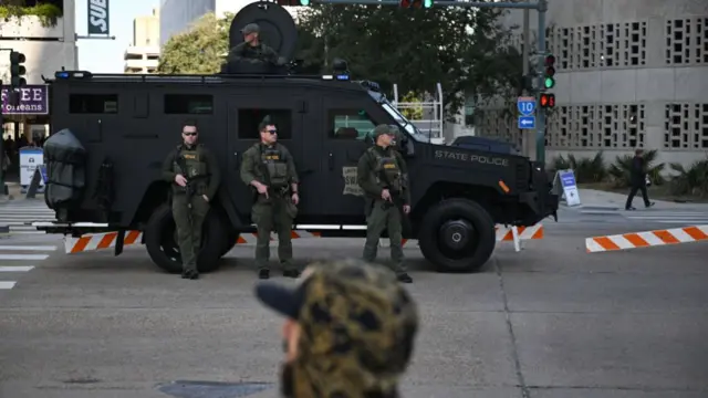 Three armed police stand in front of a matte black police van, with another armed officer coming through the roof of the van