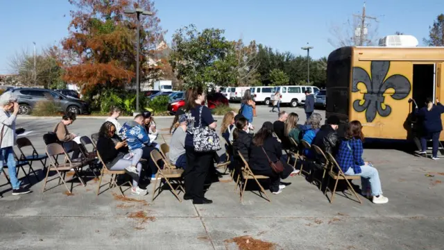 Six rows of chairs with people sitting on them in what appears to be a car park. A yellow van is pictured on the right with a woman attempting to step inside.