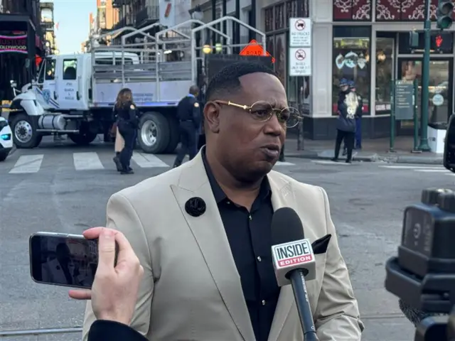 Rapper Master P in a beige jacket and black shirt speaking to media with street scene behind him