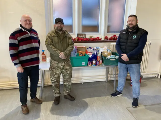 Three men  stand in a hall next to a table with donated food and items