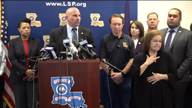 Christopher Raia, deputy assistant director of the FBI's counter-terrorism division, stands behind a podium with multiple microphones wearing a suit. He is surrounded by several people including law enforcement