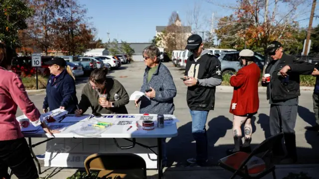 Six people line up behind a table covered with pens, paper and stickers. One person hold a cup, another a coffee cup, another two are holding their phones.