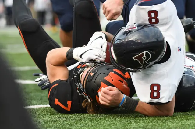 Football player in white with black helmet tackling Bech, who wears black and orange