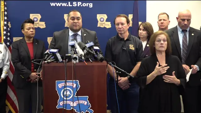 Joshua Jacson speaks behind a podium with several microphones. He is wearing a suit with a blue shirt surrounded by several people.