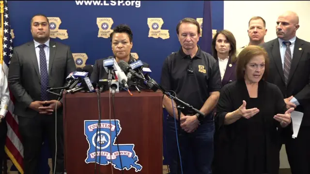 New Orleans Mayor LaToya Cantrell stands behind a podium with several microphones. She is surrounded by several people and wears a black jacket.