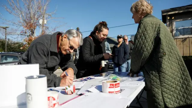 A man signs a paper in front of a queue of people. The man is wearing sunglasses and a grey jacket next to a woman wearing a black scarf and top. A woman in a green jacket stands behind the table.