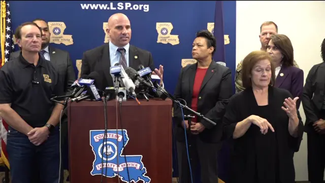 Christopher Raia stands behind podium with several microphones. He is wearing a suit with blue shirt. He stands next to Louisiana governor and others.