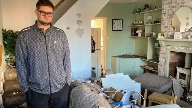 Andrew Whithers who has brown hair and beard and glasses stands in his living room assessing the flood damage