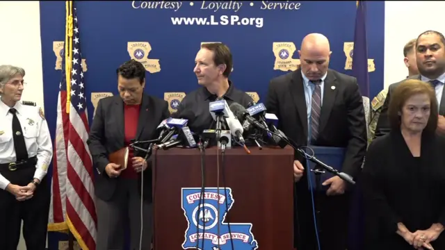 Governor of Louisiana Jeff Landry in a black top speaks at the news conference behind a podium and microphones surrounded by others.