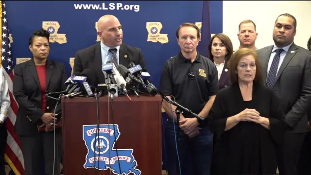 Christopher Raia speaks behind a podium with several microphones, wearing a suit, surrounded by others including law enforcement and the New Orleans mayor.