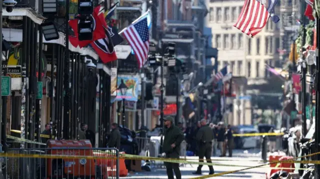 Police officers near the scene of the attack stand behind a yellow cordon. Numerous flags and signs line the sides of the street. Yellow police cordons can be seen crossing the street