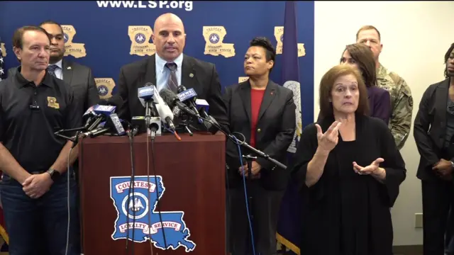 Christopher Raia stands behind podium with several microphones. He is wearing a suit with blue shirt. He stands next to Louisiana governor and others.