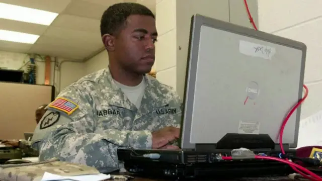 Jabbar, wearing military fatigues, sits at a desk working on a computer with a big red wire plugged into it