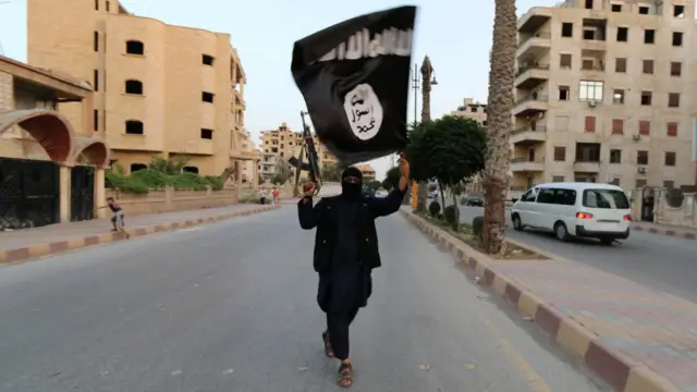 An IS member waving a black flag with white Arabic script and a circular symbol on it. He is standing in the middle of a street in Syria, pointing a Kalashikov AK-74 or AK-47 rifle in the air and dressed entirely in black - including a balaclava.