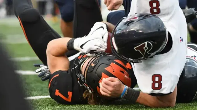 Two football players lie on the grass after a tackle. One is in a white jersey and the other in a black and orange jersey with the number seven