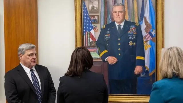 Mark Milley stands next to his newly unveiled portrait during Pentagon ceremony. Portrait hands on a white wall with a wooden panel to its right, two women - one brunette (L) one blonde (R) - have their backs to the camera watching the painting