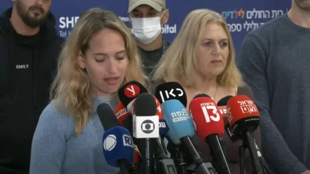 Yamit Ashkenazi addresses the press in a blue sweater as she stand behind a lectern holding several mics, her mom, in a grey, long-sleeved sweater, stands next to her