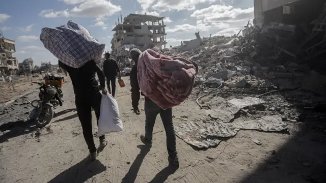 Two men carry their belongings, one in bags, the other inside a blanket. They walk amid a devastated landscape, with rubble and hollowed out buildings.