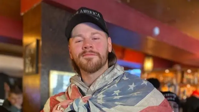 Man in brown jacket with an American flag draped across his shoulders and a "Make America Great Again" cap