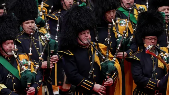 People play the bagpipes and wear fur hats while marching in uniform