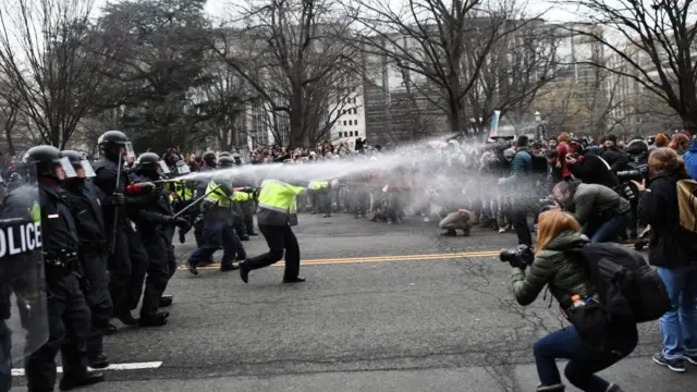 A line of protesters faces off against a line of police. Police officers are spraying something at the crowd