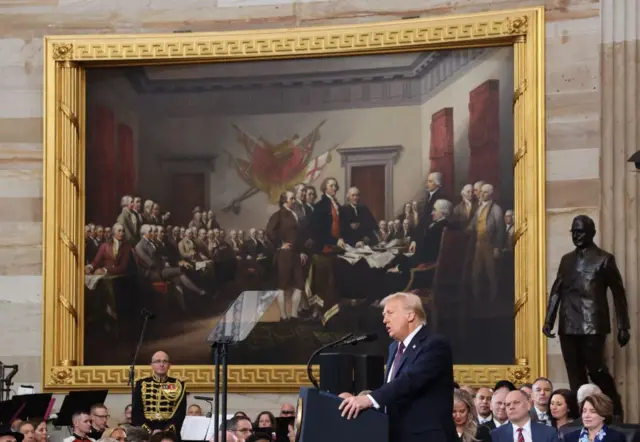 Trump speaking into a microphone. There's a massive fancy painting behind him and lots of people sat down watching on