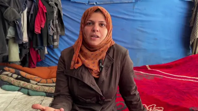 Ayat Abedrabo, a mother of nine, in her tent sitting down on the ground wearing a brown raincoat and orange patterned head scarf. Red and white blankets can be seen covering the ground with a pile of more blankets folder behind her to her right. Clothes are handing from the blue tent in the background