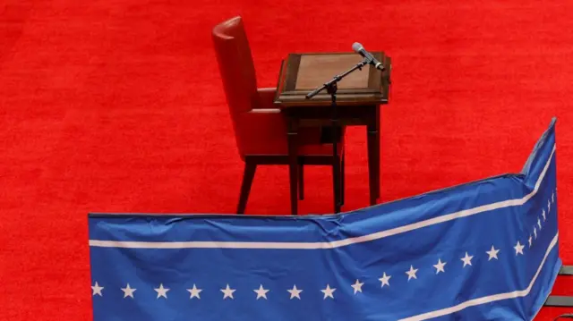 Wooden desk with red leather armchair set up on the red carpeted stage of Arena One ahead of Trump's arrival