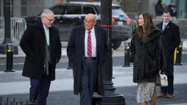 Rudy Giuliani walking along a road with a man and a woman next to him. He looks at the man. He wears a long black coat, a suit, and a tie with bright red stripes