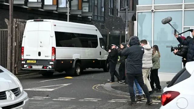 A Prison van escorted by a police van arrives at court as photographers wearing coats take photos