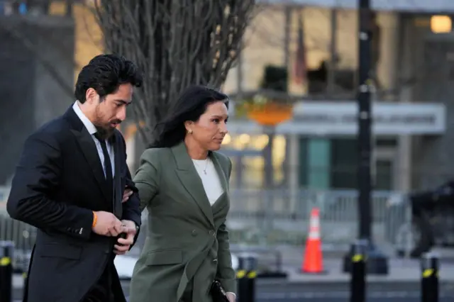 From side on, Gabbard walks holding the arm of a man next to her. She has a green blazer on and a white shirt