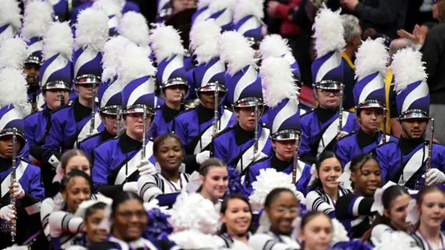 A marching band participates in a parade at the Capital One Arena. They wear purple uniforms, with some wearing tall, feathered hats.