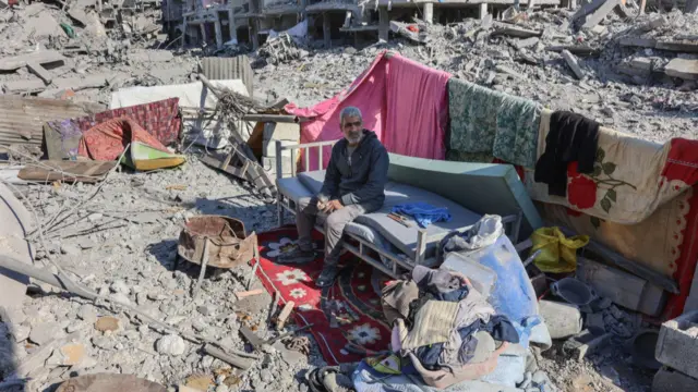 A man sits on a makeshift bed amid rubble and blankets. The landscape is devastated with bombed out buildings and rubble.