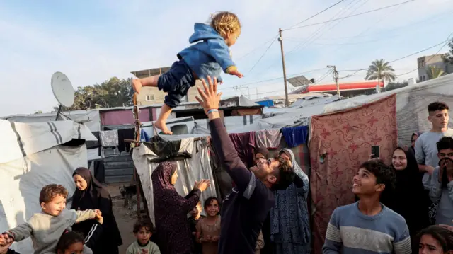 A man throws a child into the air as displaced Palestinians celebrate at a tent camp following a ceasefire