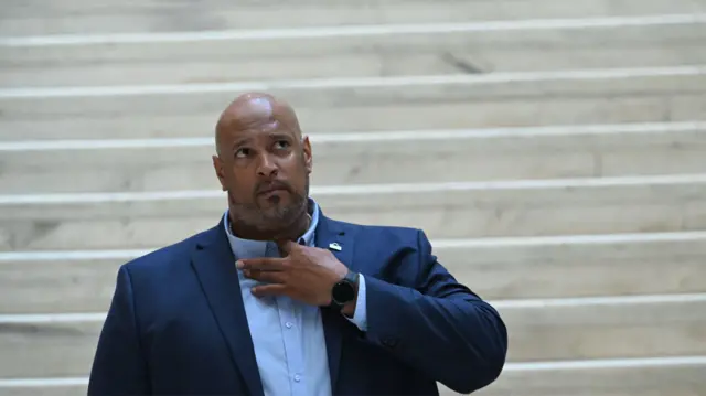 Headshot of Dunn with white marble stairs behind him. He wears a navy suit and holds one hand up to his shirt collar