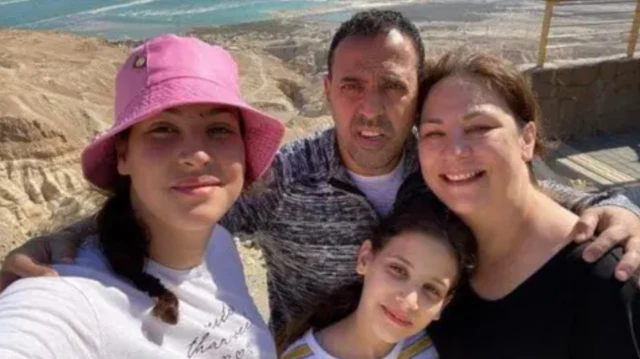 Selfie taken of family with the sea in the far distance. A man has his one arm round a woman and the other around a child. A younger child stands in front of the man in the middle of the group.