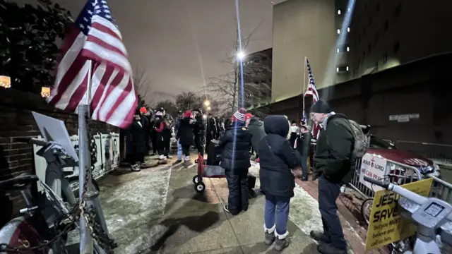 A crowd gathers outside a jail. There are American flags and placards dotted about