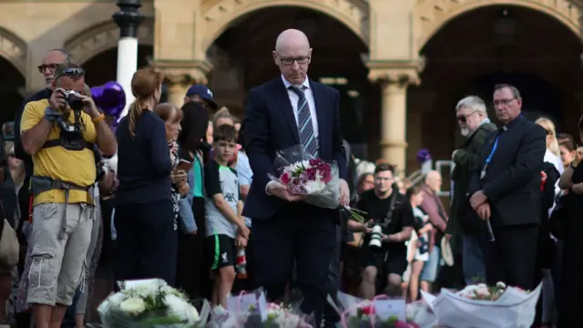 MP for Southport Patrick Hurley lays flowers, as media stand behind him