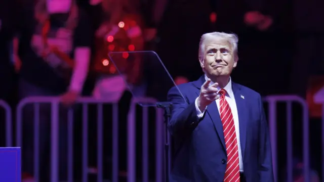 Donald Trump addresses supporters at a rally. he is pointing. he is wearing a suit and red tie.